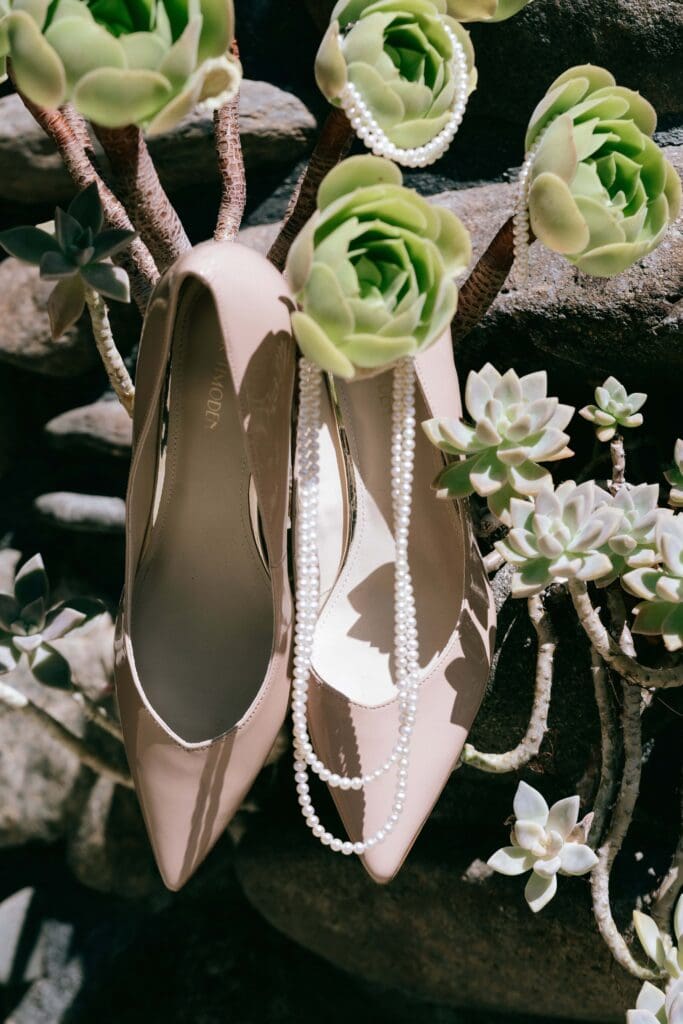 Pink nude heels hang on succulent growing out of a stone wall and pearl necklace hangs in front