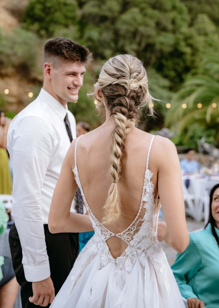 Close up of the back of the bride's dress with a low back and beaded, floral details aand long braided hair as the groom laughs in the background at their outdoor summer wedding reception