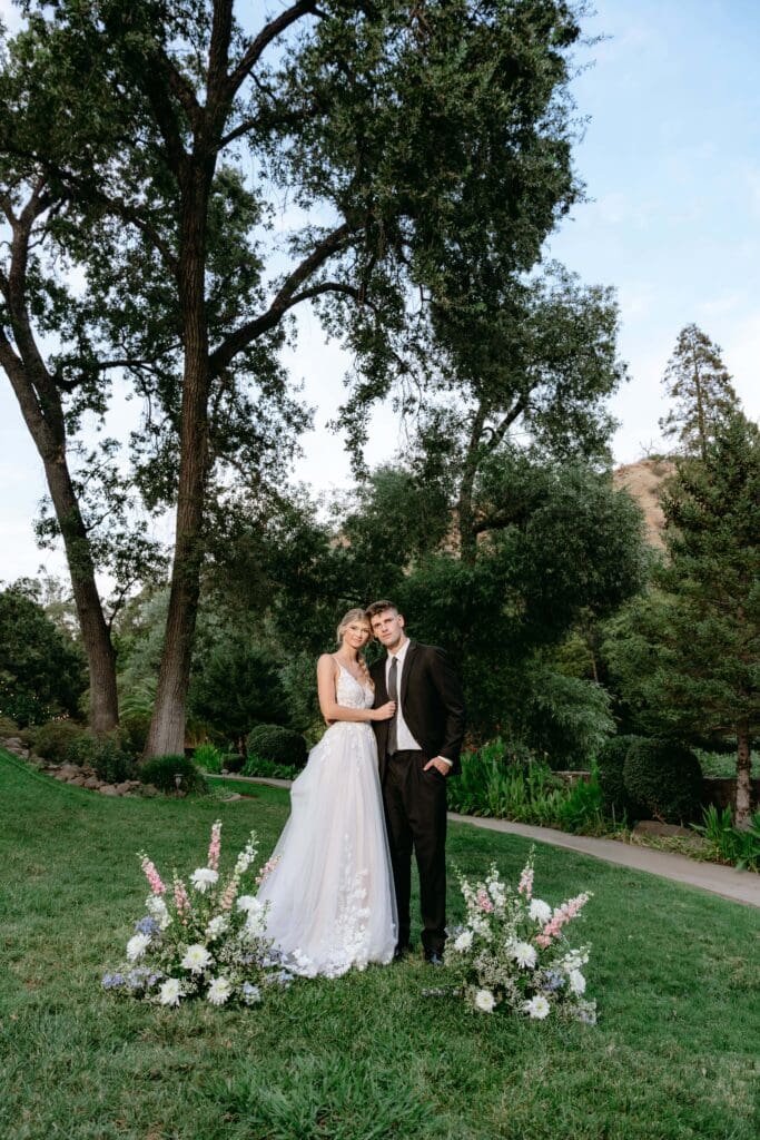 Wide portrait of Bride and groom with trees in the background and floral arrangements at their feet