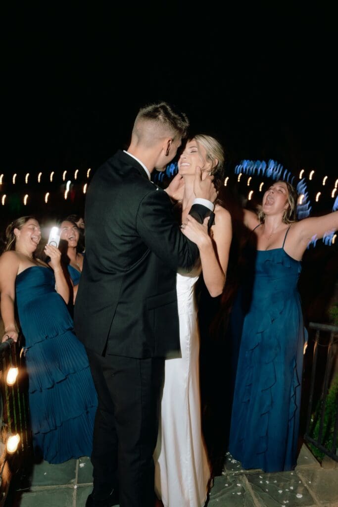 Groom is holding the bride's face, leaning in for a kiss as bridesmaids cheer in the background