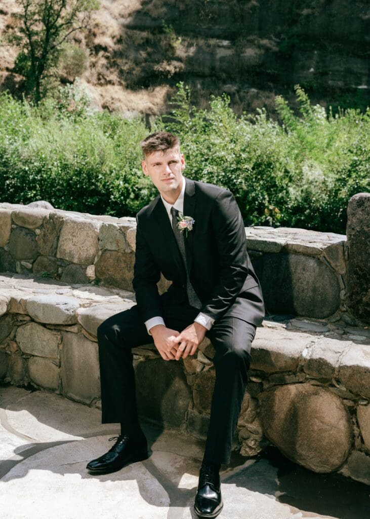 Portrait of groom looking into the camera, sitting on stone bench backlit by the sin