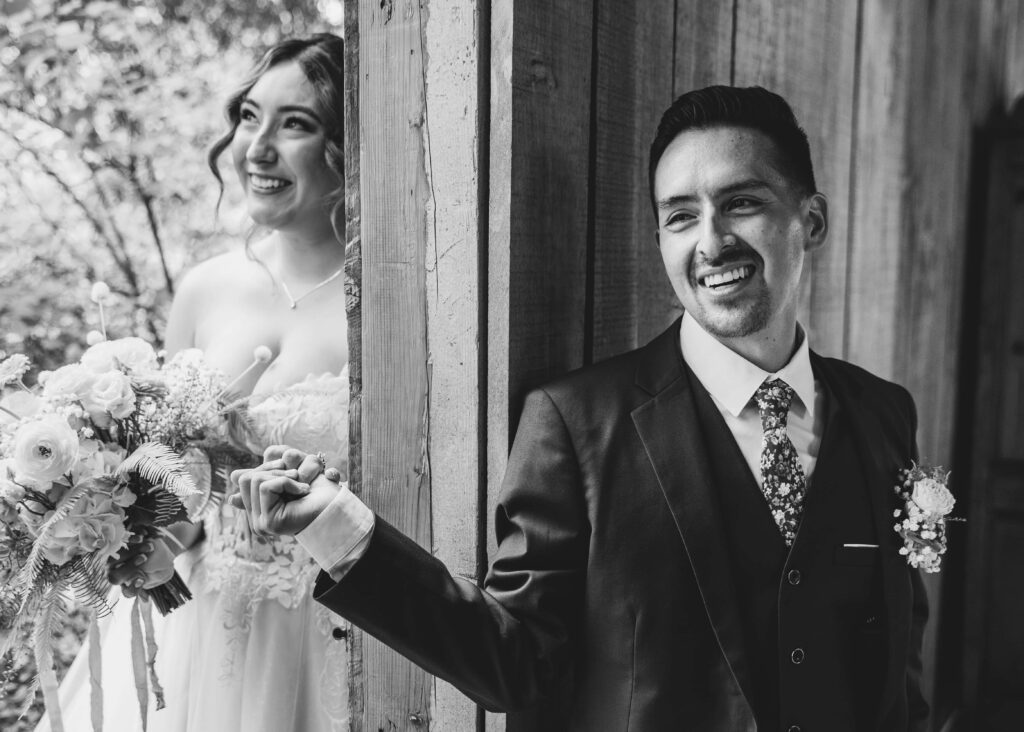 Black and white image groom holding bride's hand without seeing each other as they reach around a wall