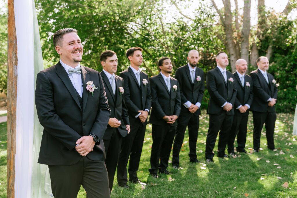 Groom smiling and standing at the alter with his groomsmen lined up beside him during the aisle reveal as he is seeing his bride walk down the aisle for the first time